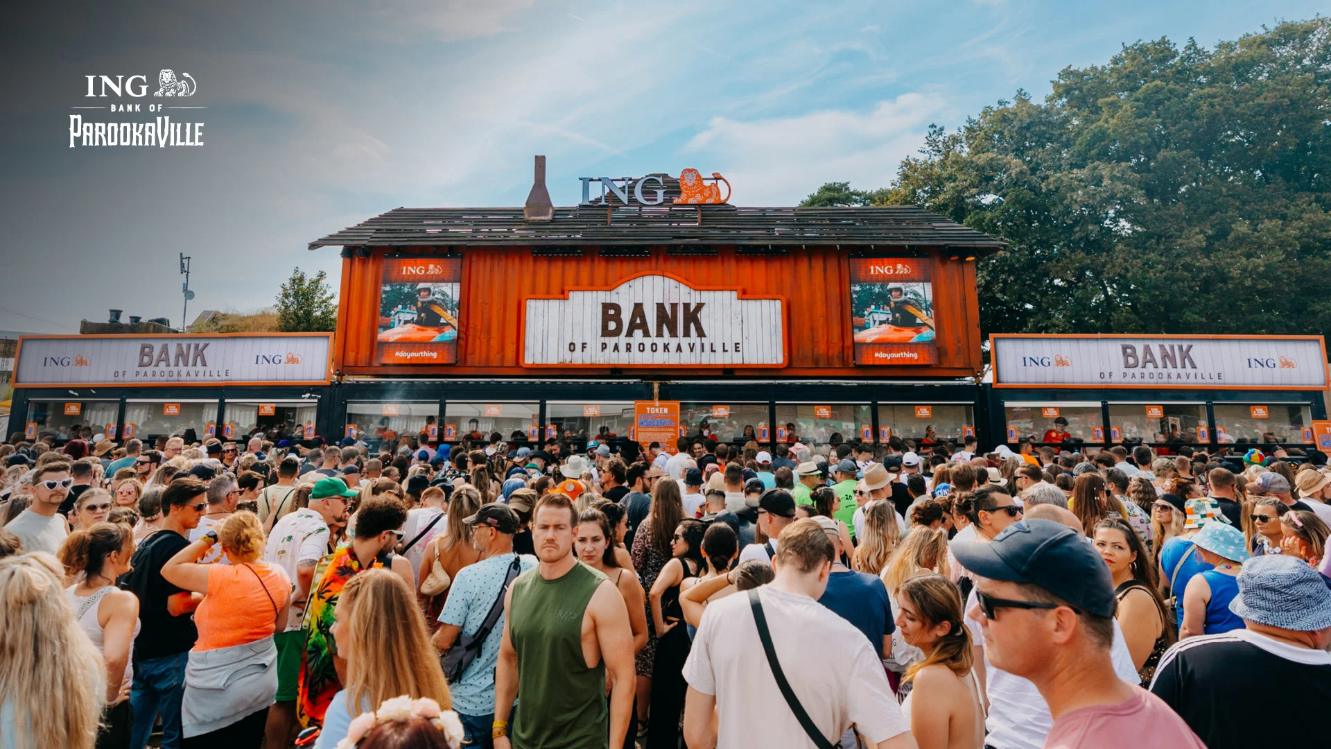 Die „ING Bank of PAROOKAVILLE“ – Festivalbesucher:innen stehen vor dem auffällig gestalteten Bankgebäude in Schiffscontainer-Optik, um Guthaben für Cashless Payment aufzuladen. Über dem Gebäude prangt das ING-Logo.