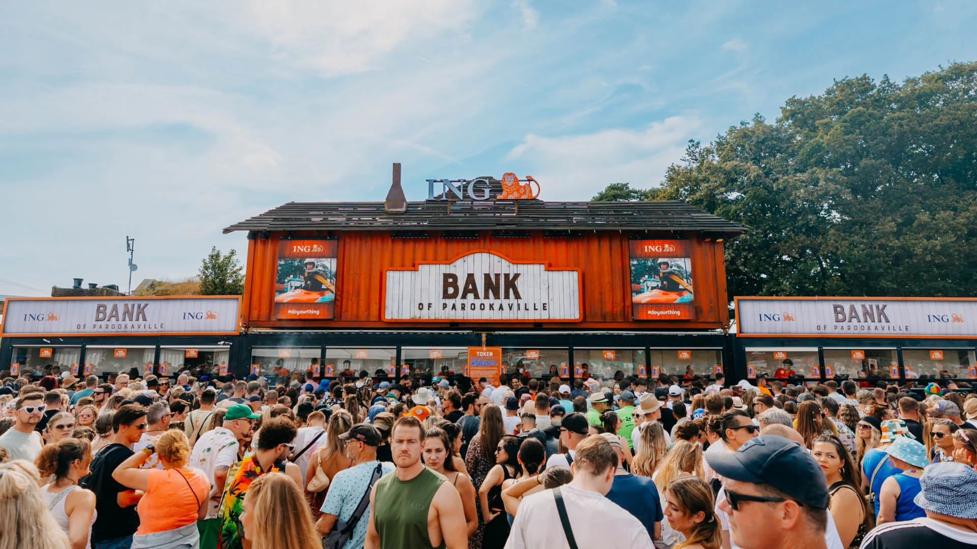 Die „ING Bank of PAROOKAVILLE“ – Festivalbesucher:innen stehen vor dem auffällig gestalteten Bankgebäude in Schiffscontainer-Optik, um Guthaben für Cashless Payment aufzuladen. Über dem Gebäude prangt das ING-Logo.