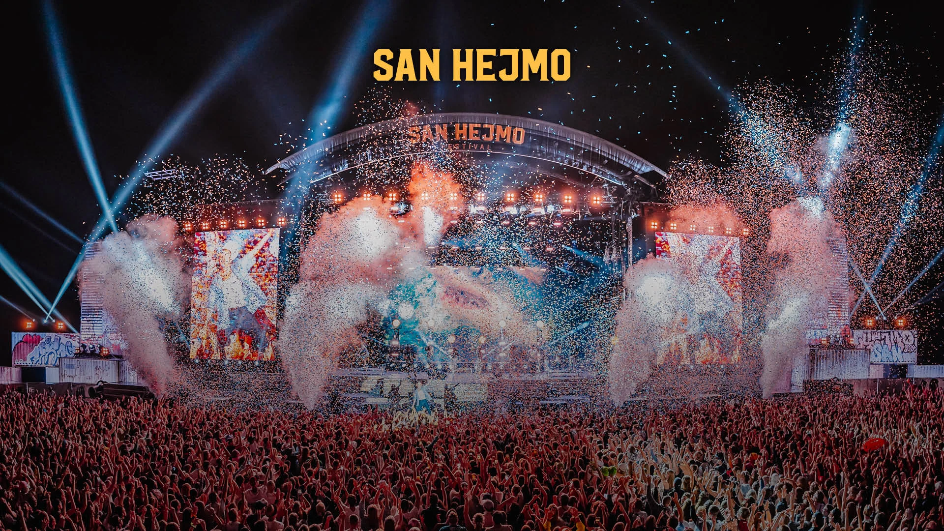 Blick auf die Hauptbühne des San Hejmo Festivals bei Nacht, mit einer großen Menschenmenge vor der Bühne, Konfettiregen, Nebeleffekten, Lichtstrahlen und LED-Leinwänden – über der Bühne leuchtet der Schriftzug „SAN HEJMO“.