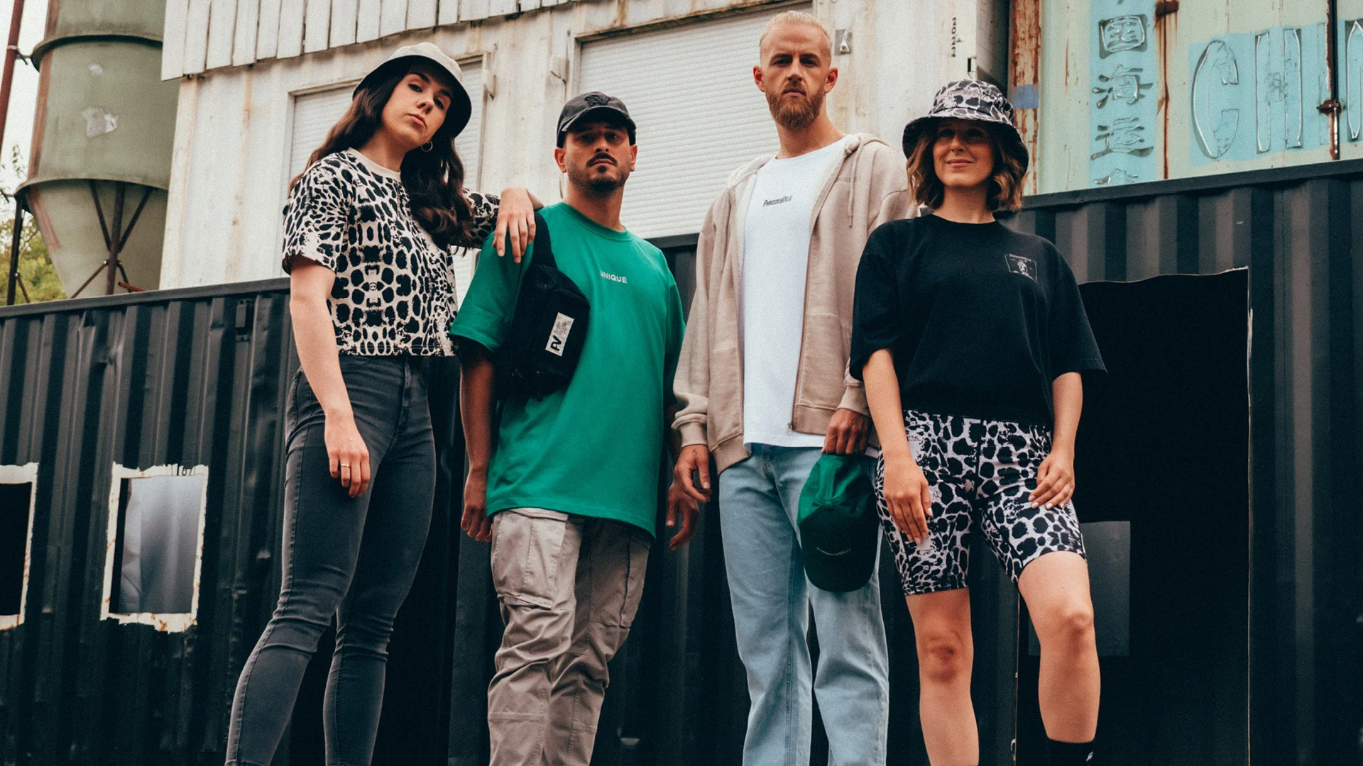 Fünf junge Erwachsene posieren in urbaner Umgebung in PAROOKAVILLE-Merchandise-Outfits. Sie tragen lässige Streetwear mit Animal-Prints, Bucket-Hats, Oversized-Shirts und Hoodies. Der Look wirkt modern, selbstbewusst und festivalbereit.